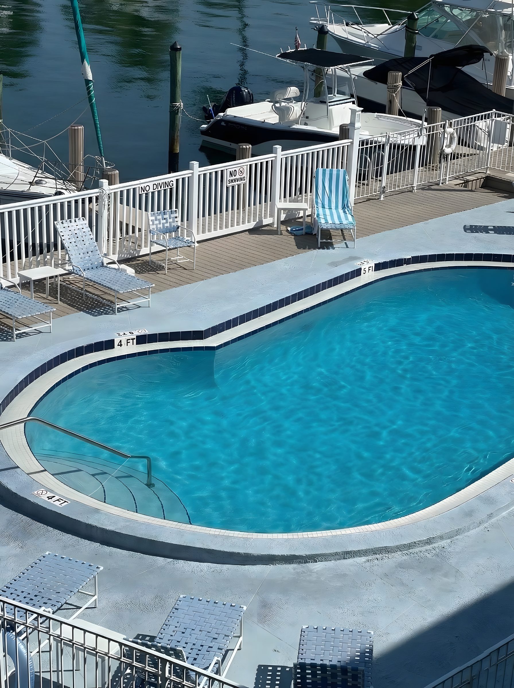 The pool seen from the deck, with loungers, the seawall, and boats moored just beyond.